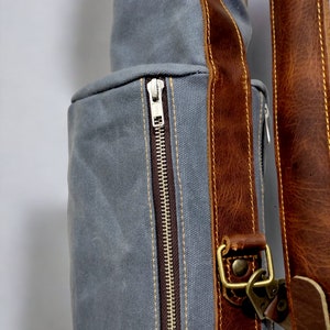 May include: A gray canvas bag with a brown leather strap and a zipper closure. The bag has a brown leather trim and a brass buckle.