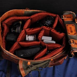 Op de afbeelding: Een bruine leren cameratas met meerdere vakken en zakken. De tas is open en gevuld met camera-apparatuur, waaronder een lens, een drone en een batterij.