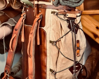 May include: A brown leather and canvas backpack with a rolled green blanket on top. The backpack has a leather strap with the initials "C.G.T." on it.