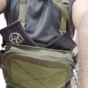 May include: A green canvas bag with a zipper closure and a velcro patch. The bag has two straps and is worn over the shoulder. Two black magazine pouches are attached to the straps.