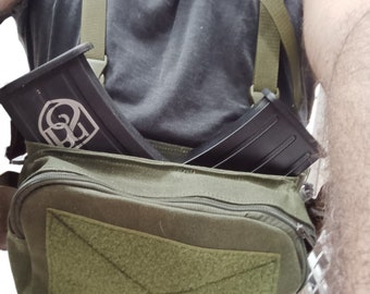 May include: A green canvas bag with a zipper closure and a velcro patch. The bag has two straps and is worn over the shoulder. Two black magazine pouches are attached to the straps.