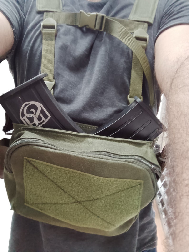 May include: A green canvas bag with a zipper closure and a velcro patch. The bag has two straps and is worn over the shoulder. Two black magazine pouches are attached to the straps.