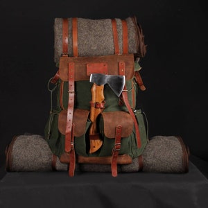 Op de afbeelding: Een groene canvas rugzak met bruine leren details, riemen en zakken. Een bijl met een houten handvat is aan de voorkant bevestigd. Een opgerolde grijze deken is aan de boven- en onderkant bevestigd. Ontworpen voor buitenactiviteiten.