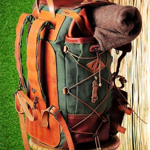 May include: A green canvas and brown leather backpack with a brown wool blanket inside. The backpack has a leather strap with a metal buckle and a leather handle. The backpack is sitting on a wooden stand.