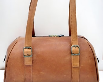 May include: A tan leather handbag with two top handles and a rectangular shape. The bag features two straps with metal buckles on the front. The bag has a zipper closure and is made of a smooth, durable leather.