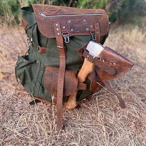 May include: A green canvas rucksack with brown leather detailing, a top flap, and several pockets. A brown leather sheath holds a small axe with a wooden handle. The rucksack has adjustable straps and metal fittings.