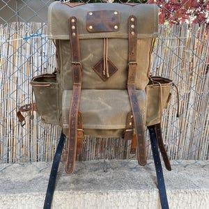 May include: A brown canvas and leather backpack with multiple straps and buckles. The backpack has two side pockets and a diamond-shaped patch on the front.