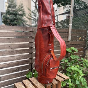 Puede incluir: Una bolsa de golf de cuero marr&oacute;n rojizo con cremallera de cuerpo entero, un bolsillo lateral con cremallera y una correa para el hombro. La bolsa est&aacute; sobre una caja de madera al aire libre.