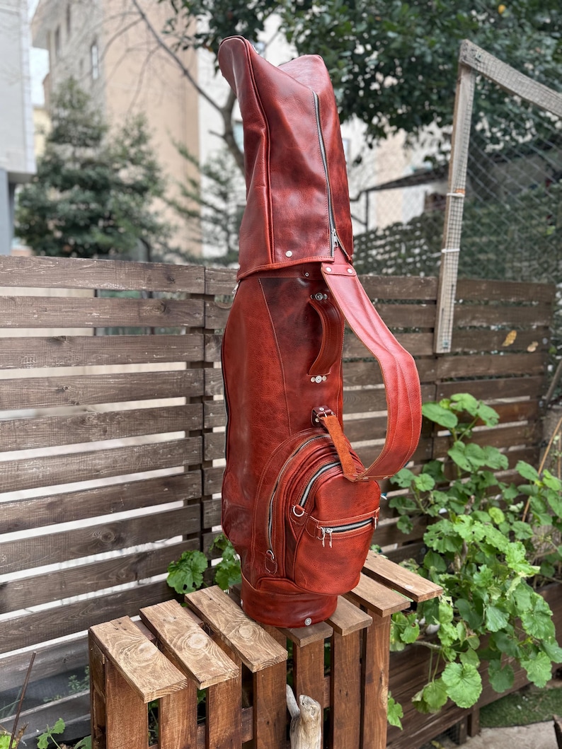 Puede incluir: Una bolsa de golf de cuero marr&oacute;n rojizo con cremallera de cuerpo entero, un bolsillo lateral con cremallera y una correa para el hombro. La bolsa est&aacute; sobre una caja de madera al aire libre.