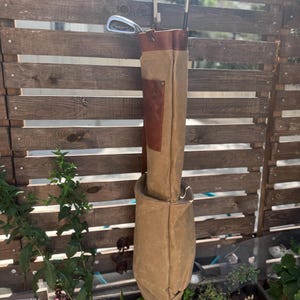 May include: A tan golf bag with brown leather detailing and a brown leather top. The bag is suspended from a wooden fence, with golf clubs inside. The bag is made of canvas material.