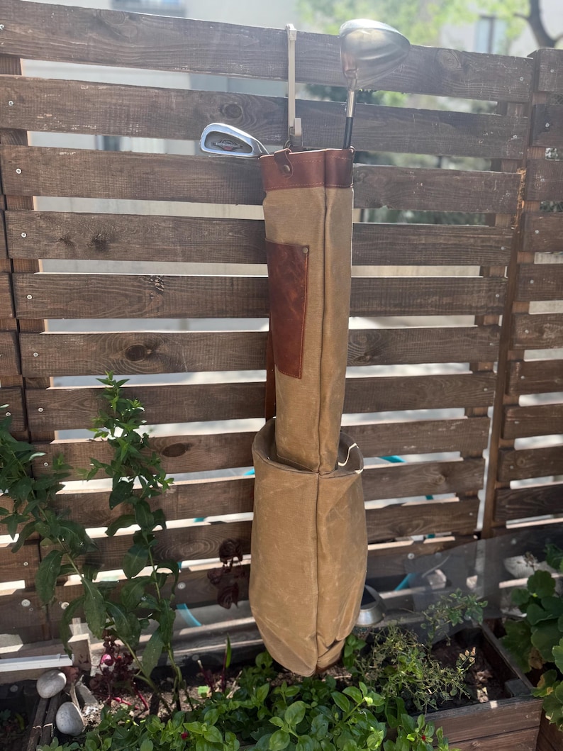 May include: A tan golf bag with brown leather detailing and a brown leather top. The bag is suspended from a wooden fence, with golf clubs inside. The bag is made of canvas material.