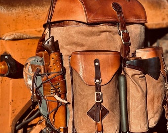 May include: A brown leather and canvas backpack with multiple pockets and straps. The backpack has a large main compartment and a smaller front pocket. There is a knife and a sheath attached to the side of the backpack. The backpack is sitting on a rusty orange surface.