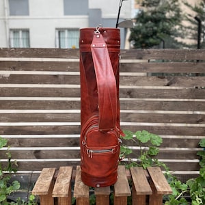 Puede incluir: Una bolsa de golf de cuero marr&oacute;n rojizo con una correa para el hombro y un bolsillo lateral con cremallera. Se ven dos palos de golf que sobresalen de la parte superior de la bolsa. La bolsa es cil&iacute;ndrica y est&aacute; sobre un pal&eacute; de madera.