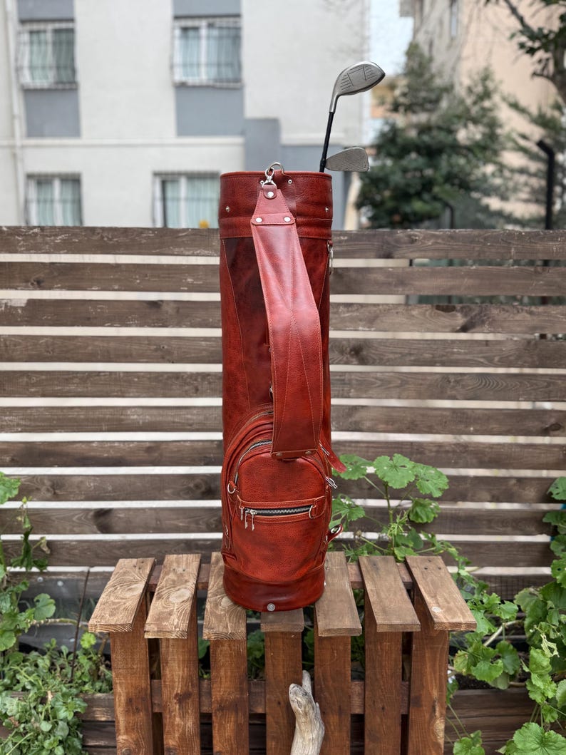 Puede incluir: Una bolsa de golf de cuero marr&oacute;n rojizo con una correa para el hombro y un bolsillo lateral con cremallera. Se ven dos palos de golf que sobresalen de la parte superior de la bolsa. La bolsa es cil&iacute;ndrica y est&aacute; sobre un pal&eacute; de madera.