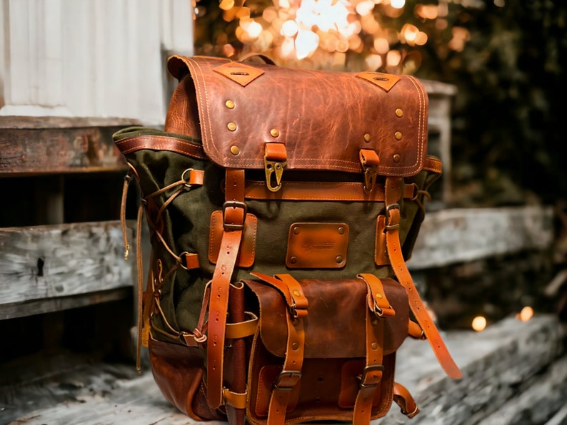May include: A brown leather and green canvas backpack with multiple straps and buckles. The backpack has a large flap with a leather patch that says "The Pack" on it.