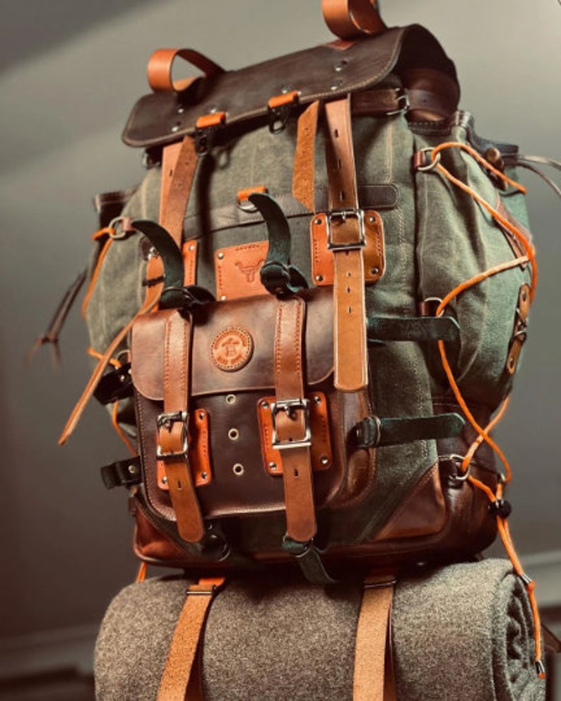 May include: A large, brown leather and canvas backpack with multiple straps and buckles. The backpack has a large front pocket with a flap and buckle closure.