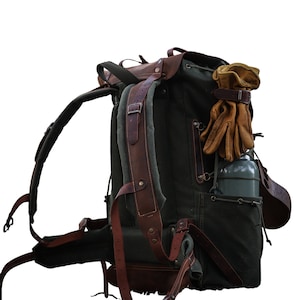 May include: A large, green canvas backpack with brown leather straps and buckles. The backpack has a side pocket and a water bottle holder. A pair of brown leather gloves are attached to the top of the backpack.