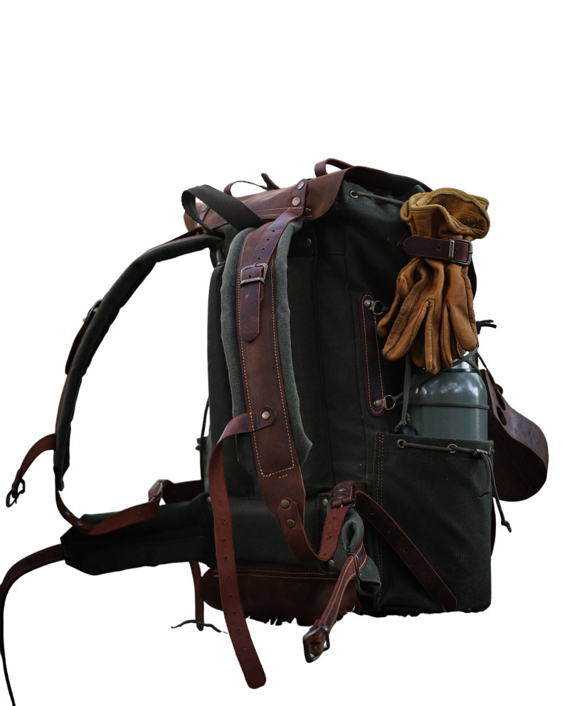 May include: A large, green canvas backpack with brown leather straps and buckles. The backpack has a side pocket and a water bottle holder. A pair of brown leather gloves are attached to the top of the backpack.