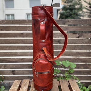 Puede incluir: Una bolsa de golf de cuero marr&oacute;n rojizo con un bolsillo lateral y una correa para el hombro a juego. Se ven dos palos de golf. La bolsa est&aacute; sobre un banco de madera contra una valla de madera.