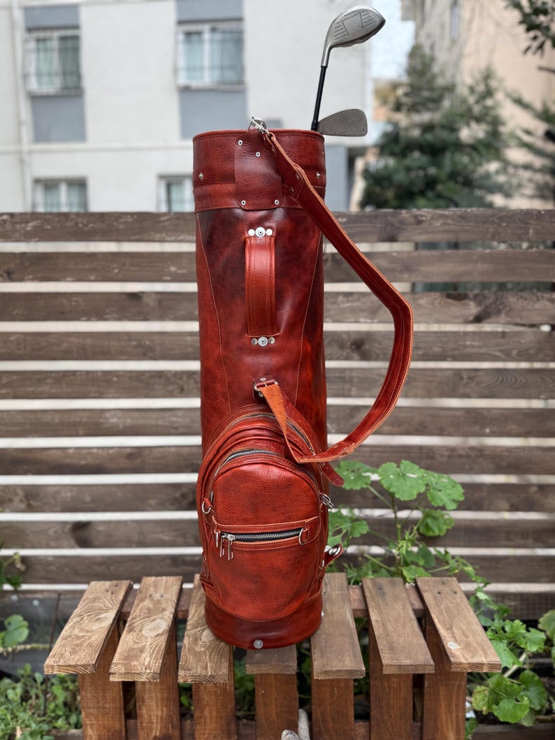 Puede incluir: Una bolsa de golf de cuero marr&oacute;n rojizo con un bolsillo lateral y una correa para el hombro a juego. Se ven dos palos de golf. La bolsa est&aacute; sobre un banco de madera contra una valla de madera.