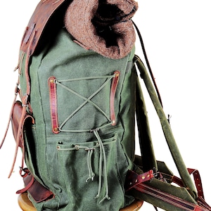 May include: A green canvas backpack with brown leather straps and accents. The backpack has a large main compartment and several smaller pockets. It is sitting on a wooden pedestal.