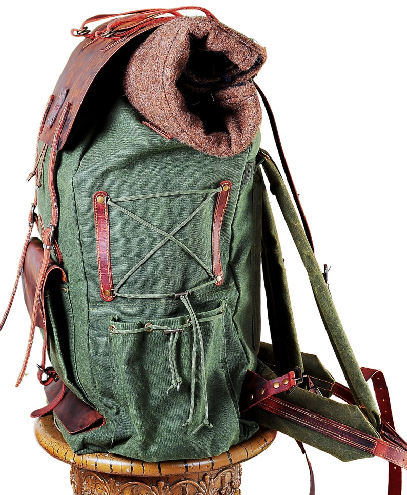 May include: A green canvas backpack with brown leather straps and accents. The backpack has a large main compartment and several smaller pockets. It is sitting on a wooden pedestal.