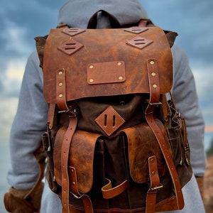 May include: A brown leather backpack with a canvas front and multiple pockets. The backpack has a distressed look and features a leather patch with the text "The Wanderer" on the front.