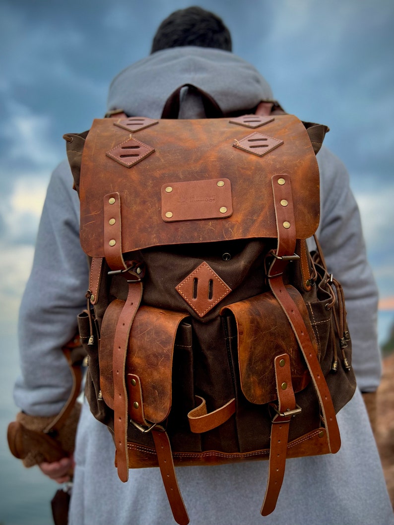 May include: A brown leather backpack with a canvas front and multiple pockets. The backpack has a distressed look and features a leather patch with the text "The Wanderer" on the front.