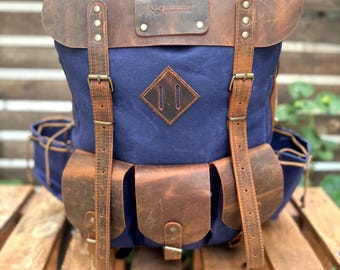 May include: A blue and brown backpack with leather accents. The backpack features a leather flap, straps, and pockets. The front of the backpack has a diamond-shaped leather patch. The text "LIFE OF CHIEF HANDMADE" is visible on a leather patch.