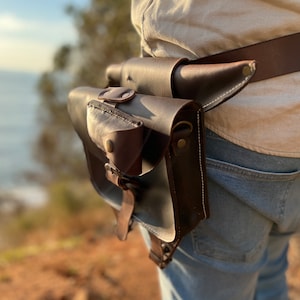 K&ouml;nnte beinhalten: Ein brauner Lederg&uuml;rtel mit einer braunen Ledertasche. Die Tasche hat eine Klappe mit einem Druckknopfverschluss und ein kleines Fach auf der Vorderseite. Der G&uuml;rtel wird um die Taille einer Person getragen, die eine blaue Jeans tr&auml;gt.