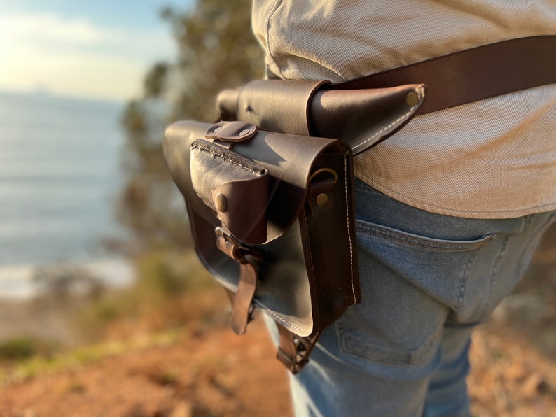 K&ouml;nnte beinhalten: Ein brauner Lederg&uuml;rtel mit einer braunen Ledertasche. Die Tasche hat eine Klappe mit einem Druckknopfverschluss und ein kleines Fach auf der Vorderseite. Der G&uuml;rtel wird um die Taille einer Person getragen, die eine blaue Jeans tr&auml;gt.