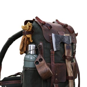 May include: A green canvas backpack with brown leather straps and buckles. The backpack has a variety of accessories attached to it, including a metal water bottle, a wooden cup, a pair of leather gloves, and an axe.