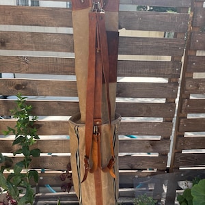 May include: A tan and brown golf bag with leather straps and accents. The bag has a cylindrical shape with a zippered pocket and a brown leather base. Golf clubs are visible inside the bag. The bag is hanging on a wooden fence.