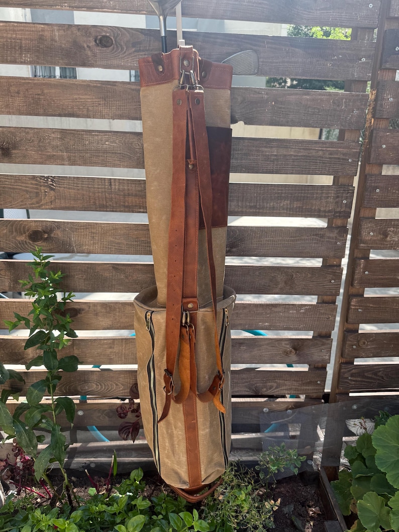 May include: A tan and brown golf bag with leather straps and accents. The bag has a cylindrical shape with a zippered pocket and a brown leather base. Golf clubs are visible inside the bag. The bag is hanging on a wooden fence.