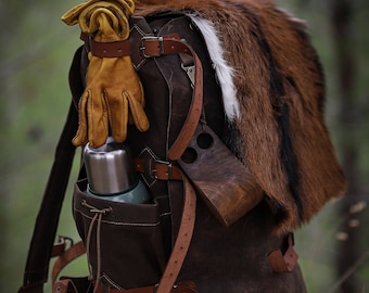 May include: A brown leather backpack with multiple straps and buckles. The backpack has a fur flap on the top and a metal thermos in the front pocket. The backpack is sitting on a bed of pine needles.