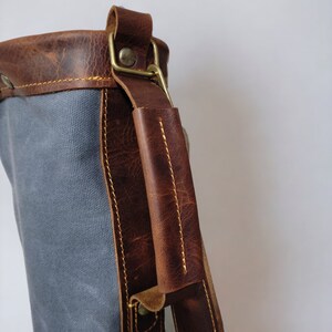 May include: A gray canvas bag with a brown leather strap and handle. The bag has a brass buckle and a brass rivet on the strap.