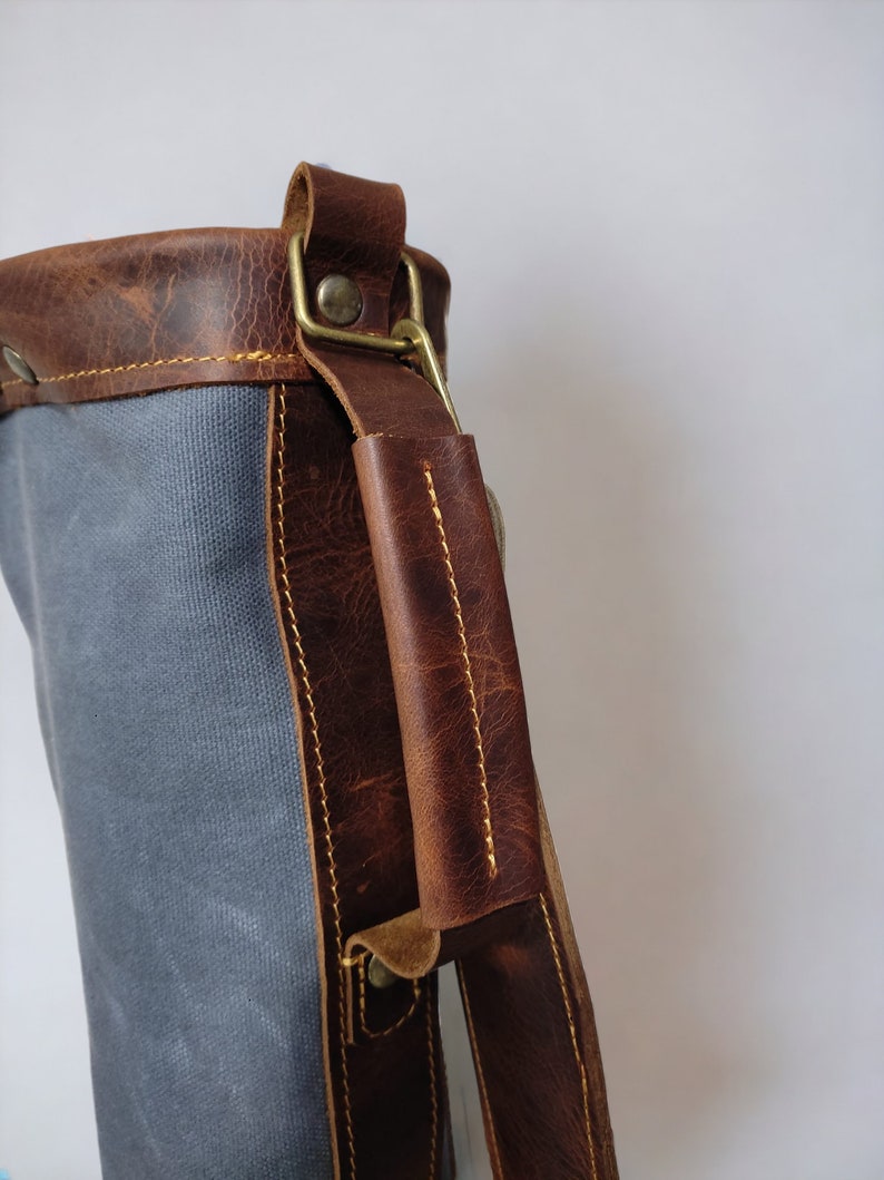 May include: A gray canvas bag with a brown leather strap and handle. The bag has a brass buckle and a brass rivet on the strap.
