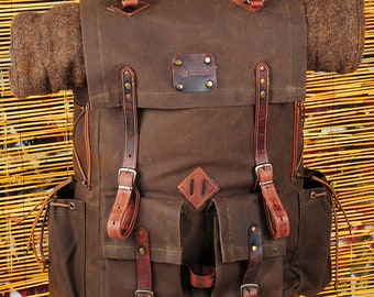 May include: A brown canvas backpack with leather straps and buckles. The backpack has a large main compartment and two smaller front pockets. The backpack is sitting on a wooden stand.