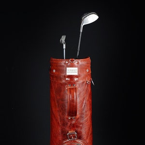 Może przedstawiać: Brązowa sk&oacute;rzana torba golfowa z dwoma kijami golfowymi. Torba ma uchwyt i klamrę. Tekst "99 PERCENT HANDMADE" znajduje się na prostokątnej metalowej płytce na torbie.