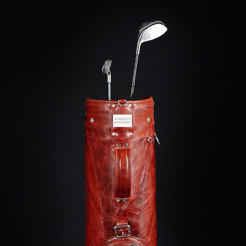 Może przedstawiać: Brązowa sk&oacute;rzana torba golfowa z dwoma kijami golfowymi. Torba ma uchwyt i klamrę. Tekst "99 PERCENT HANDMADE" znajduje się na prostokątnej metalowej płytce na torbie.