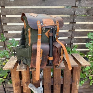May include: A green and brown leather backpack with multiple pockets and straps. The backpack has a flap closure and is accented with tan leather straps and details. It is sitting on a wooden pallet.