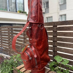 Puede incluir: Una bolsa de golf de cuero marr&oacute;n rojizo con un compartimento principal grande y m&uacute;ltiples bolsillos con cremallera. La bolsa tiene una correa para el hombro y un asa para llevarla. La bolsa mide aproximadamente 91 cm de alto.
