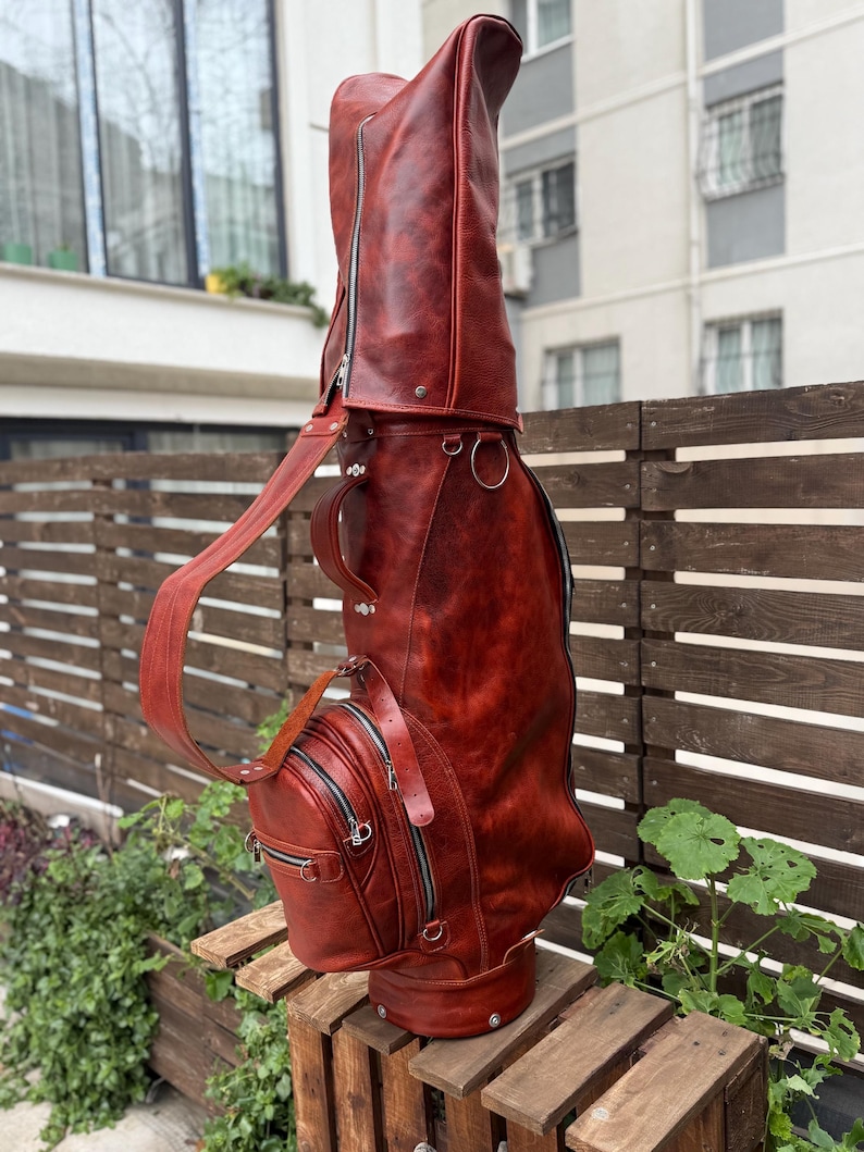 Puede incluir: Una bolsa de golf de cuero marr&oacute;n rojizo con un compartimento principal grande y m&uacute;ltiples bolsillos con cremallera. La bolsa tiene una correa para el hombro y un asa para llevarla. La bolsa mide aproximadamente 91 cm de alto.