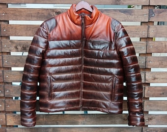May include: A brown leather puffer jacket with a high collar and a zip-up front. The jacket has a two-tone design, with a lighter brown color on the shoulders and collar. The jacket has a quilted pattern and two front pockets.