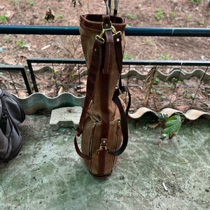 May include: A brown leather golf bag with a canvas exterior and multiple pockets. The bag is hanging on a metal railing and has several golf clubs inside.