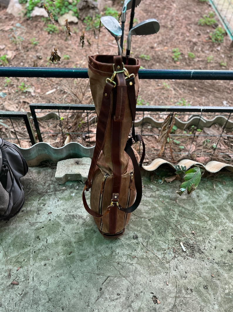 May include: A brown leather golf bag with a canvas exterior and multiple pockets. The bag is hanging on a metal railing and has several golf clubs inside.