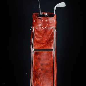 Może przedstawiać: Wysoki, czerwonobrązowy sk&oacute;rzany worek na kije golfowe z dwoma kijami. Torba ma teksturowany wygląd, ze srebrnymi zamkami błyskawicznymi i nitami. Kije są srebrno-czarne, na czarnym tle.