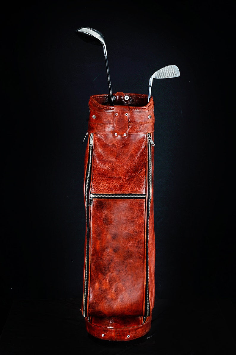 Może przedstawiać: Wysoki, czerwonobrązowy sk&oacute;rzany worek na kije golfowe z dwoma kijami. Torba ma teksturowany wygląd, ze srebrnymi zamkami błyskawicznymi i nitami. Kije są srebrno-czarne, na czarnym tle.