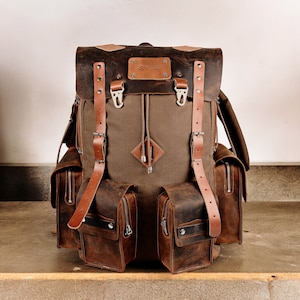 Pode incluir: Uma mochila de couro marrom com vários bolsos e alças. A mochila tem um painel frontal de lona com um remendo em forma de diamante.