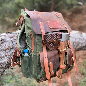 Puede incluir: Una mochila de lona verde con detalles y correas de cuero marrón. La mochila tiene un bolsillo grande en la parte delantera con un bolsillo más pequeño en el lateral. Un pequeño hacha está unida al lateral de la mochila.